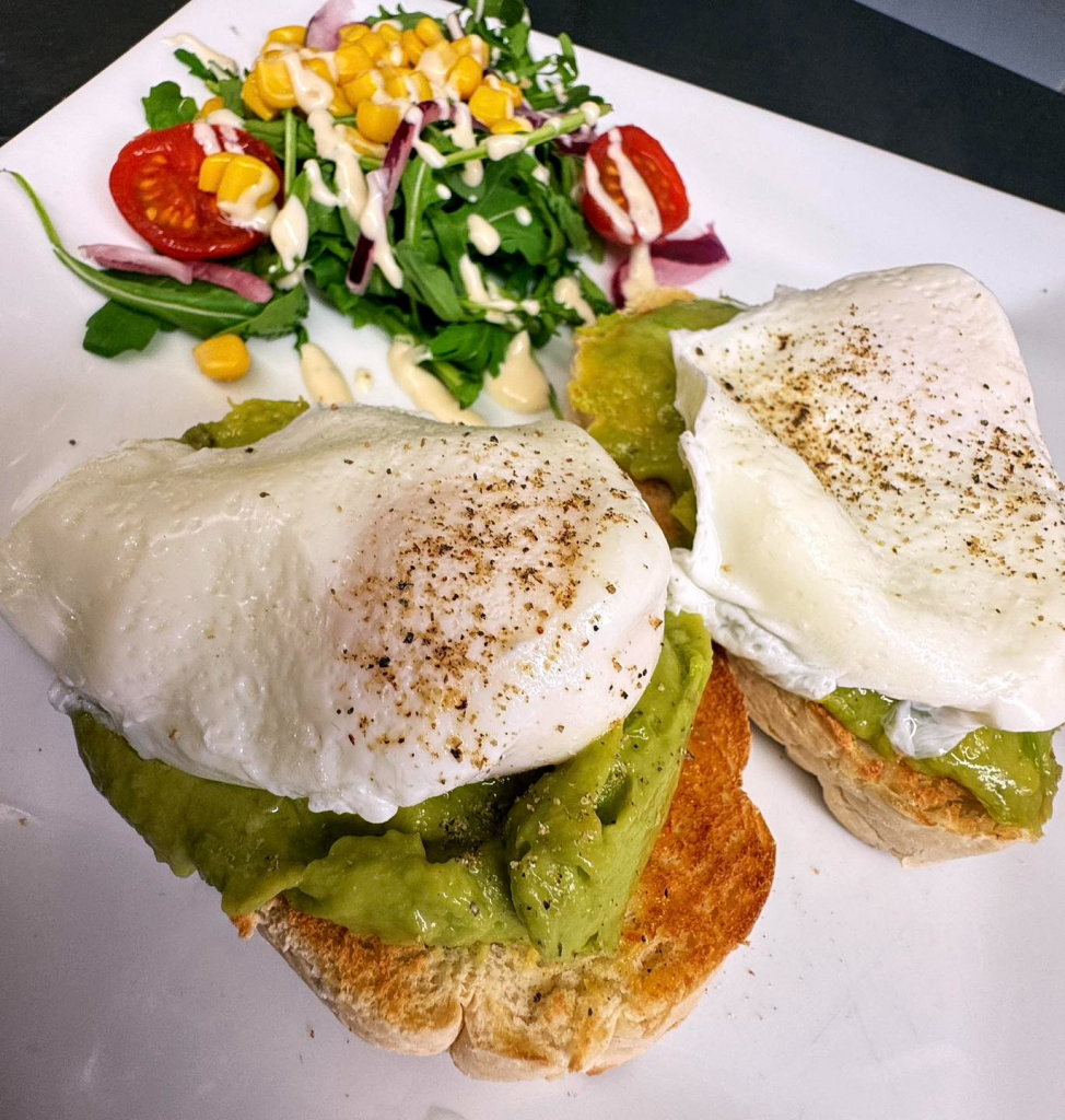 Poached eggs on avocado toast with side salad, featuring cherry tomatoes, sweetcorn, red onion, rocket, and dressing