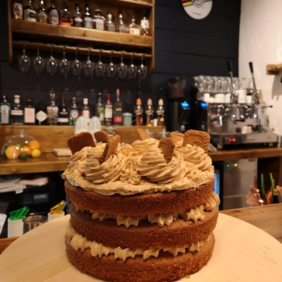 Three-layer cake with biscuit toppings on a wooden countertop in a cosy café setting with beverages in the background