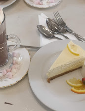 Slice of lemon cheesecake on a white plate with citrus slices, accompanied by a hot chocolate with marshmallows