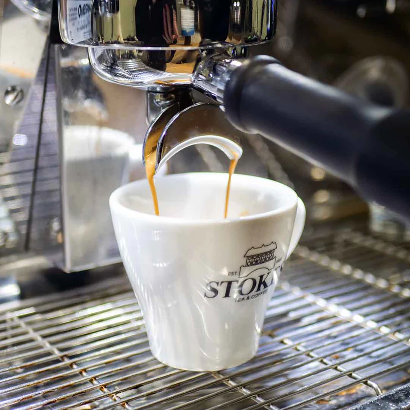 Espresso flowing from coffee machine into a Stokes cup on a metal grate, showing brewing process in a café setting