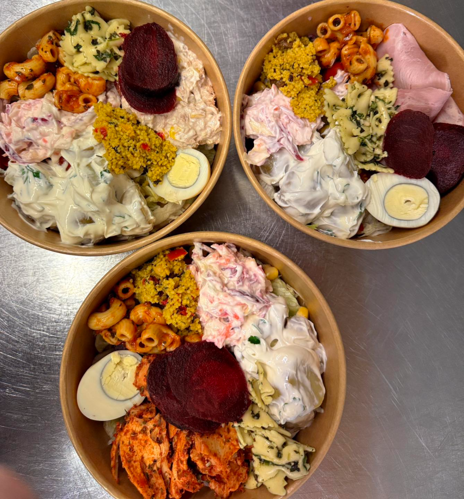 Three diverse salad bowls with pasta, beetroot, boiled eggs, couscous, ham, and creamy dressing on a metal surface