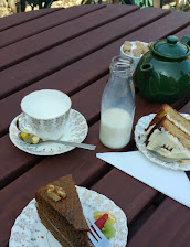 Afternoon tea with cake slices on a wooden table including a teapot, milk bottle, and sugar bowl outdoors