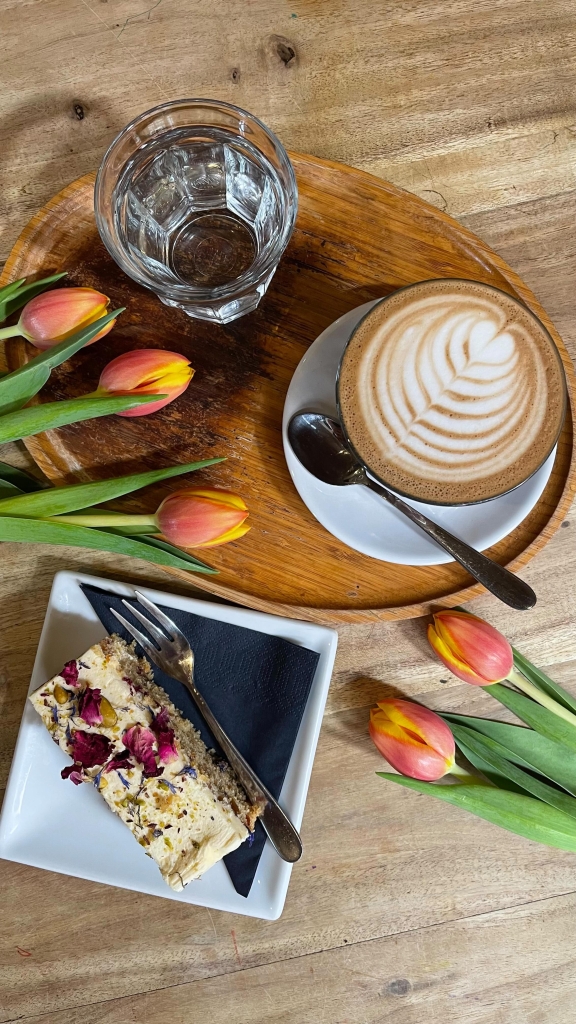 Cappuccino with latte art, floral cake slice, and tulips on a rustic wooden table setting