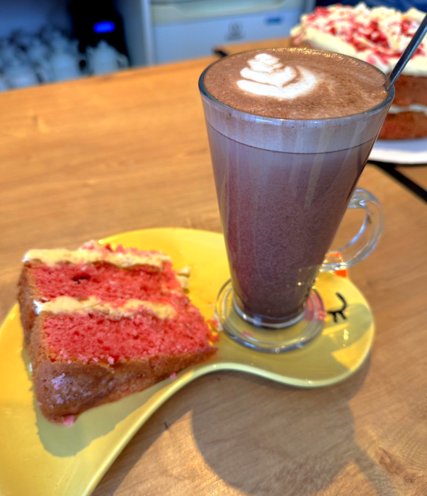 Hot chocolate and slice of pink sponge cake with cream on a yellow platter in a cosy cafe setting