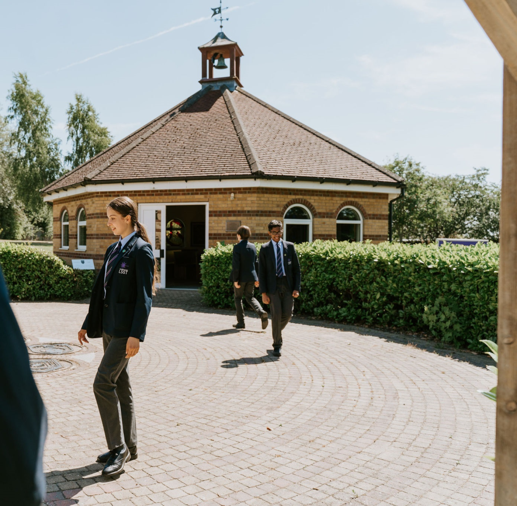 Students in school uniforms walking near a brick building on a sunny day with greenery in the background