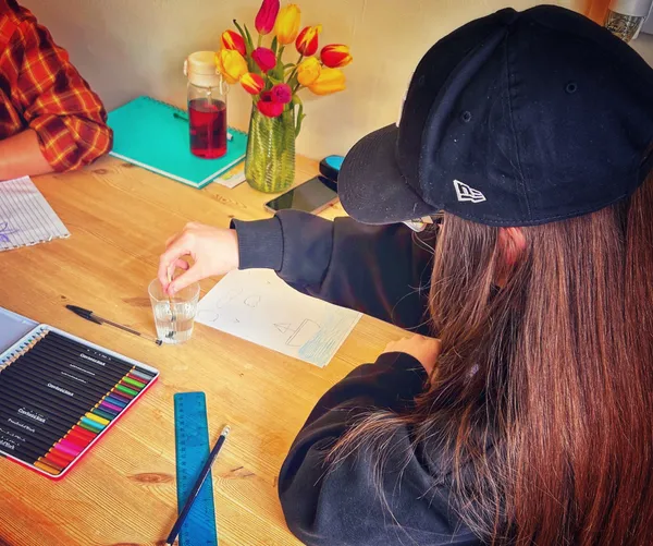 Person in cap drawing with coloured pencils at wooden table, vase of tulips, notebook and glass of water nearby