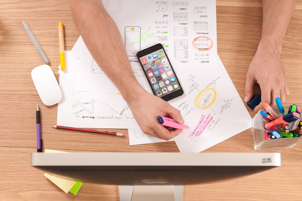 Person planning app design with smartphone, notepads, and colourful markers on wooden desk