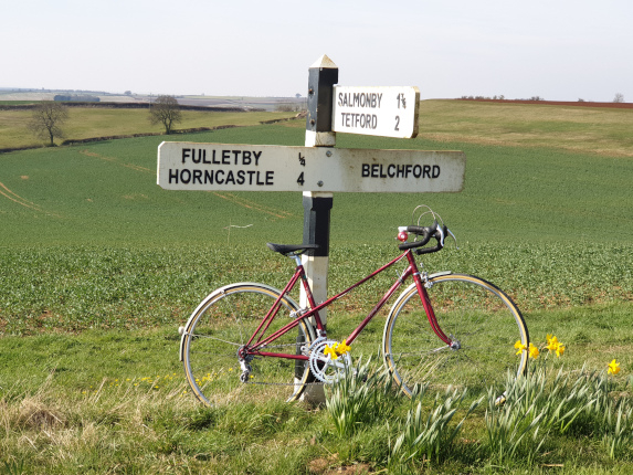 Vintage red bicycle leaning on countryside signpost in green field with daffodils in Lincolnshire, UK