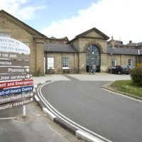 Entrance of a UK hospital featuring a directional sign and parking area on a clear day