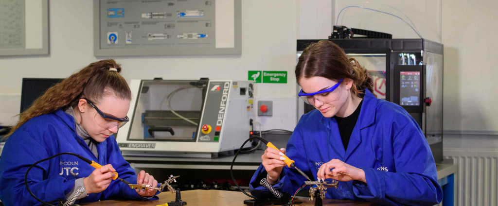 Women in blue lab coats practising soldering skills in an engineering workshop
