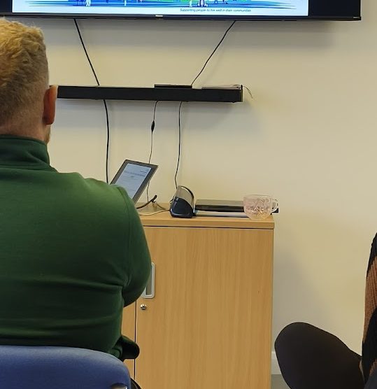 Man in green jumper attending a presentation in an office setting with a screen and laptop