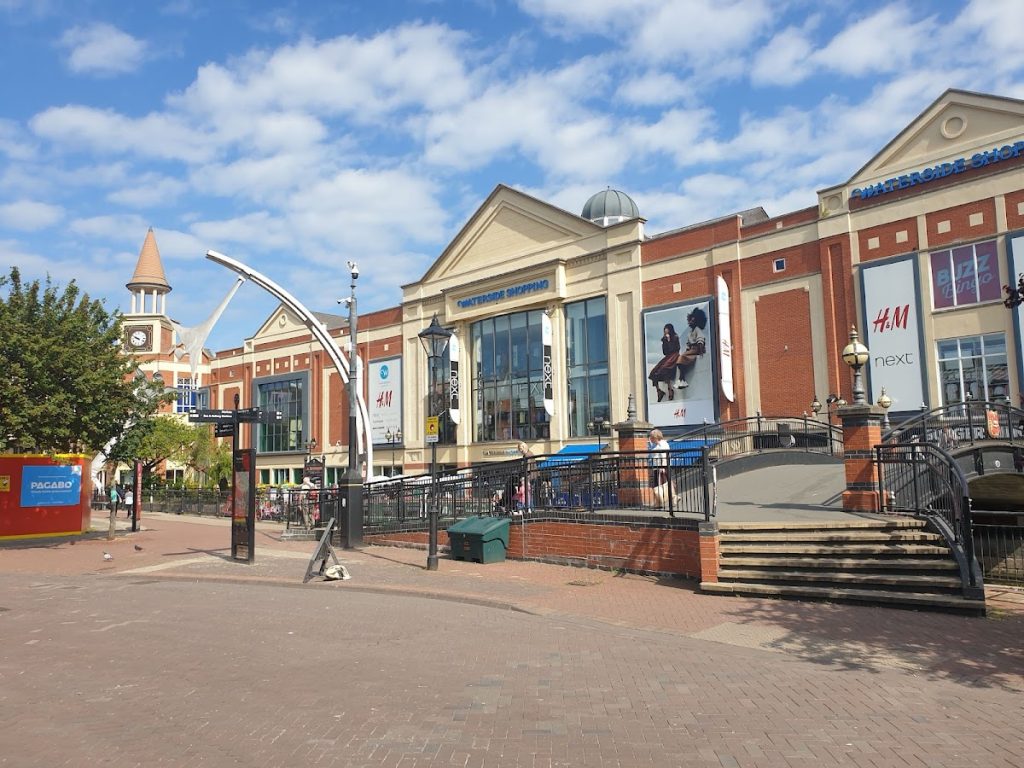 Modern shopping centre exterior with clear blue sky, featuring large retail stores and a clock tower in the background