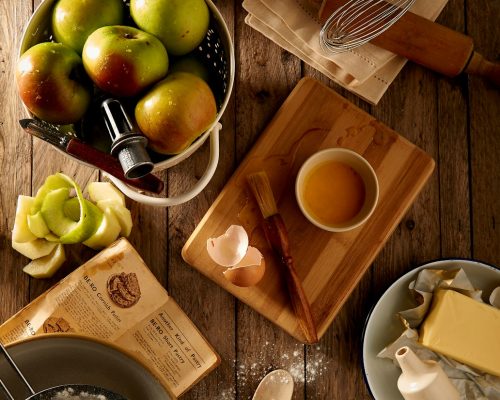 Baking ingredients and tools on a rustic wooden table with apples, flour, butter, eggs, and a whisk for apple pie preparation