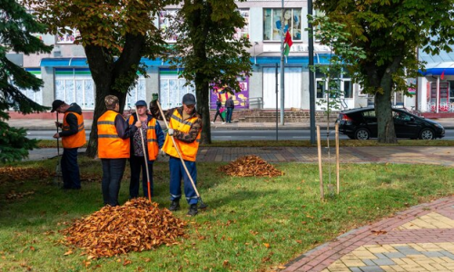 Workers in orange vests gather leaves in an urban park, showcasing community upkeep and autumn maintenance work