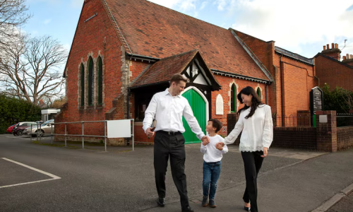 Family walking hand in hand past a traditional red brick church with green doors on a bright day