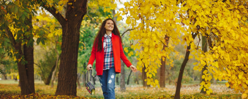 Woman strolling in vibrant autumn park with yellow leaves and trees, wearing a red jacket and carrying a small handbag