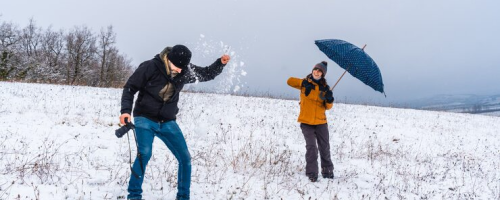 Two people having fun in a snowy field, one throwing snow, the other with an umbrella, winter landscape in the background